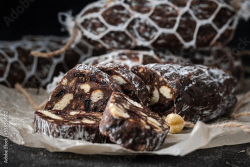 Chocolate salami filled with almonds, hazelnuts and raisins on the black background. Italian sweet sausage shaped dessert sprinkled with powdered sugar. Christmas and New Year festive pastry
