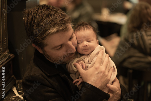 indoors lifestyle portrait of happy father holding his newborn baby girl only a few weeks old sitting on cafe taking care of the little daughter proud and cheerful © TheVisualsYouNeed