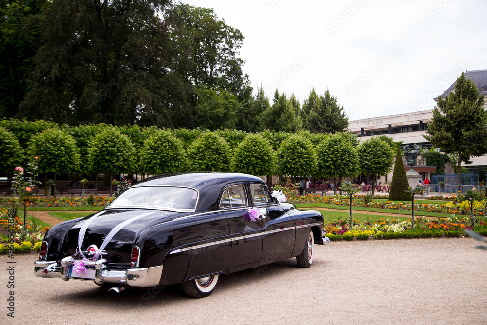 The vintage car, black and shiny, is parked in the courtyard of a cathedral in Europe. The car is decorated with flowers and ribbons.
