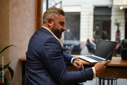 Modern senior businessman sitting in cafe and using laptop and smartphone