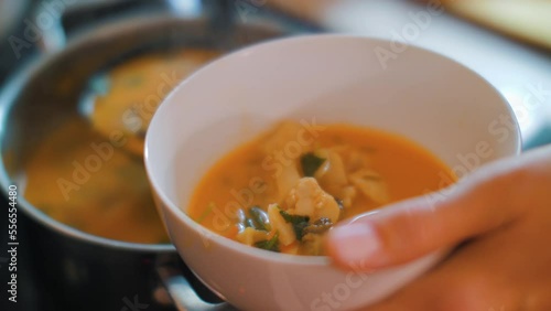 Woman pours bright orange soup with meat and vegetables in white porcelain bowl. Tasty and spicy Asian Tom Yam soup.