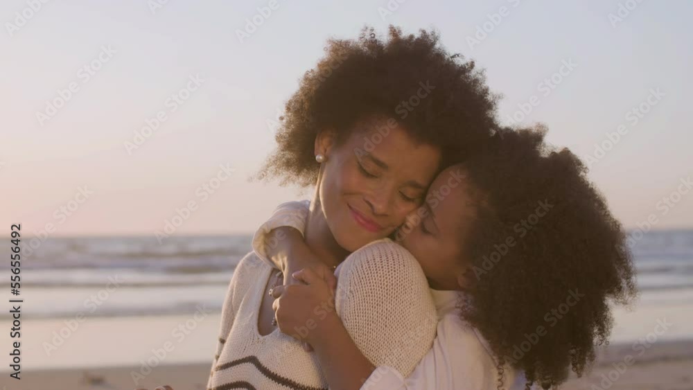 Closeup of happy mum and daughter having picnic at seashore at sunset. Cute Black girl hugging mother from behind and kissing her cheek gently. Slow motion. Childhood, mother and daughter concept. Closeup of happy mum and daughter having picnic at seashore at sunset. Cute Black girl hugging mother from behind and kissing her cheek gently. Slow motion. Childhood, mother and daughter concept.