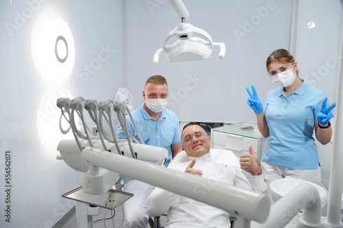 Three joyful and contented patient  doctor  dental assistant thumb up successful dental treatment remove tartar in dental office masks and gloves in blue color transparent glasses in clean stomatology