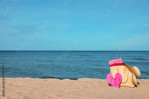 Summer bag with slippers  beach towel and straw hat on sand near sea  space for text