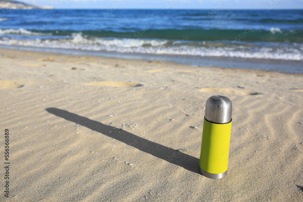 Yellow thermos with hot drink on sand near sea, space for text Yellow thermos with hot drink on sand near sea, space for text