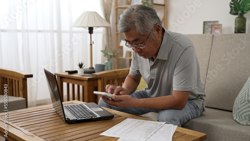 Retirement financial planning concept. senior asian man counting on calculator with family budget sitting in living room sofa using laptop computer and paper indoors