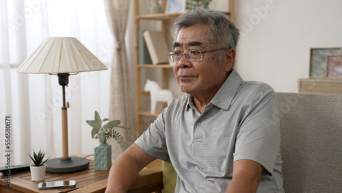 Dreamy inspired senior hoary asian man resting on comfortable sofa in morning. serious old male in eyeglasses look front while sitting on couch at home during quarantine.