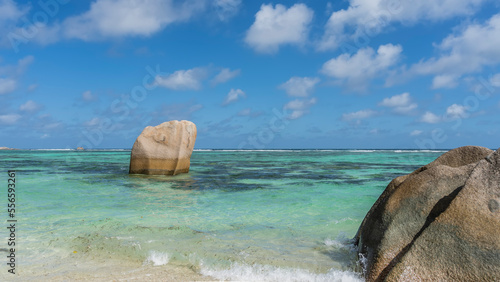 Picturesque granite boulders in the turquoise ocean. Rounded shapes, folded slopes. Waves are foaming on a sandy beach. Clouds in the blue sky. Seychelles. La Digue. Anse Source D’Argent beach 