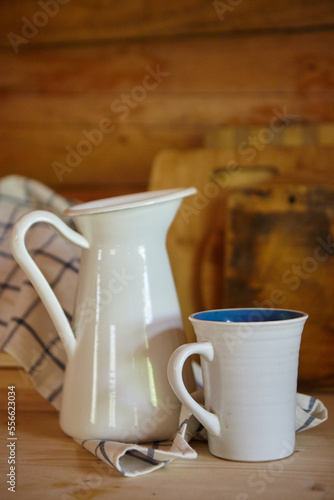 Rustic kitchen items. Enameled metal white jug for milk or water and a mug on the kitchen table in a log house