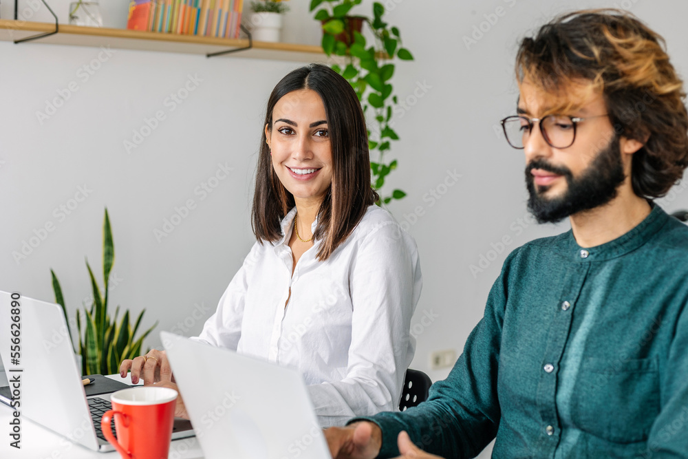 Smiling young businesswoman by colleague using laptop at desk Smiling young businesswoman by colleague using laptop at desk
