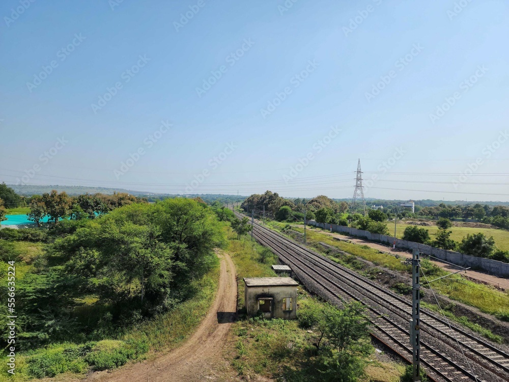 Stock photo of railway track or train pathway surrounded by green trees and plants. Electric poles and small old cabin near the railway track. Picture captured under bright sunlight at Gulbarga, India