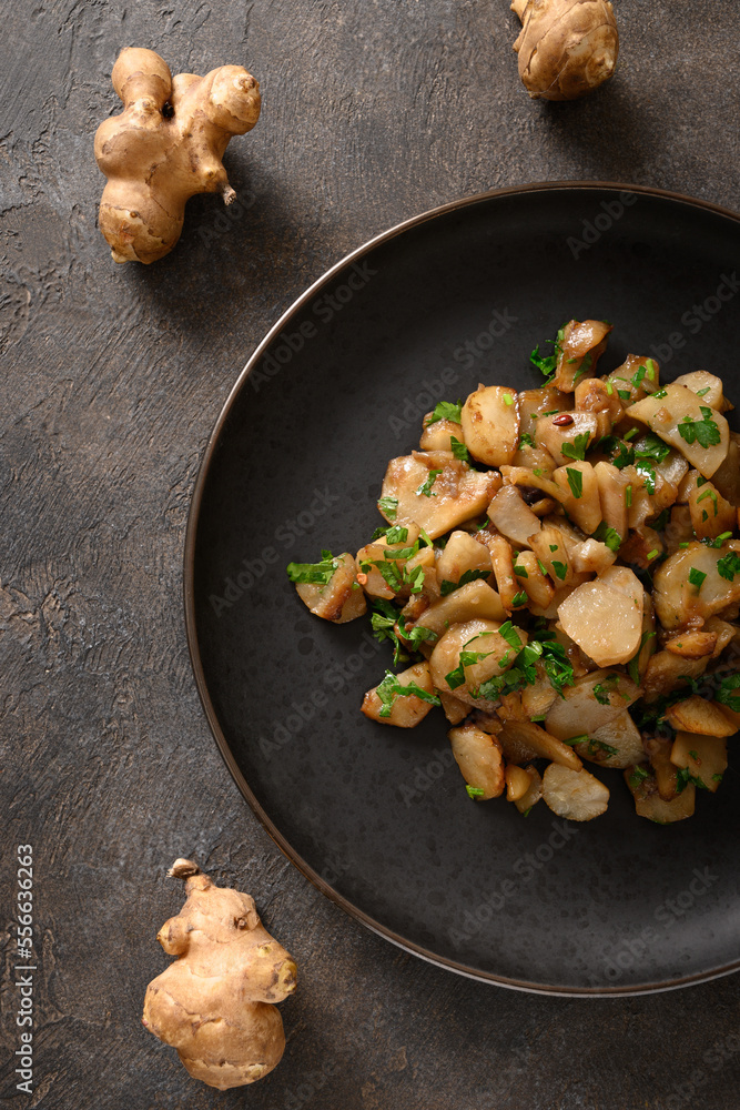 Fototapeta premium Roasted Jerusalem artichoke root, topinambur, sunchokes with garlic and parsley on brown background. Top view. Vertical format.