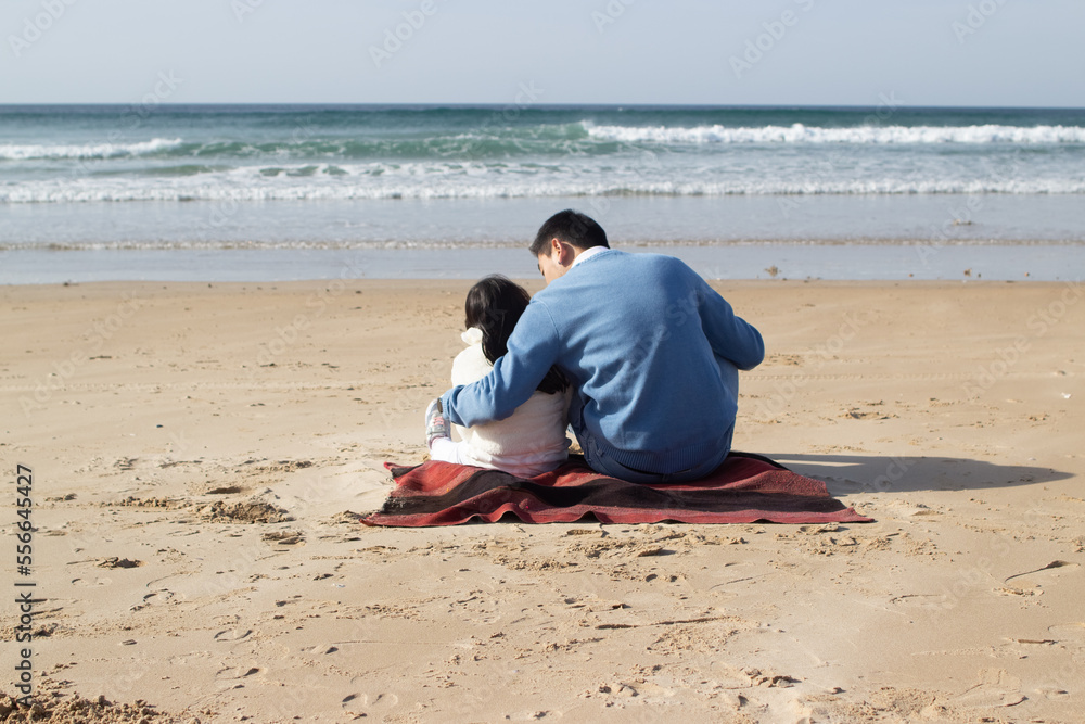 Pleased Japanese family on blanket on beach. Father and daughter in casual clothes sitting on blanket, hugging. Family, relaxation, nature concept