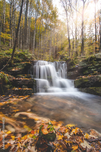 Vlasky River with waterfall illuminated with sun in an untouched landscape in the middle of an autumn mixed forest in red-orange colour. Moravka, Beskydy mountains, Czech republic