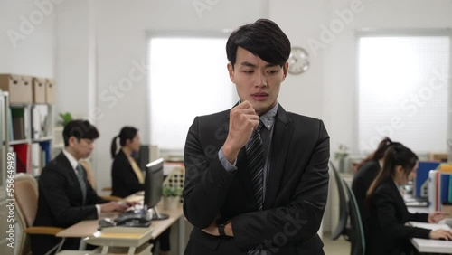 portrait of a worried asian male manager feeling anxiety about making mistake and rubbing fingers while thinking about solution. other coworkers are working at background