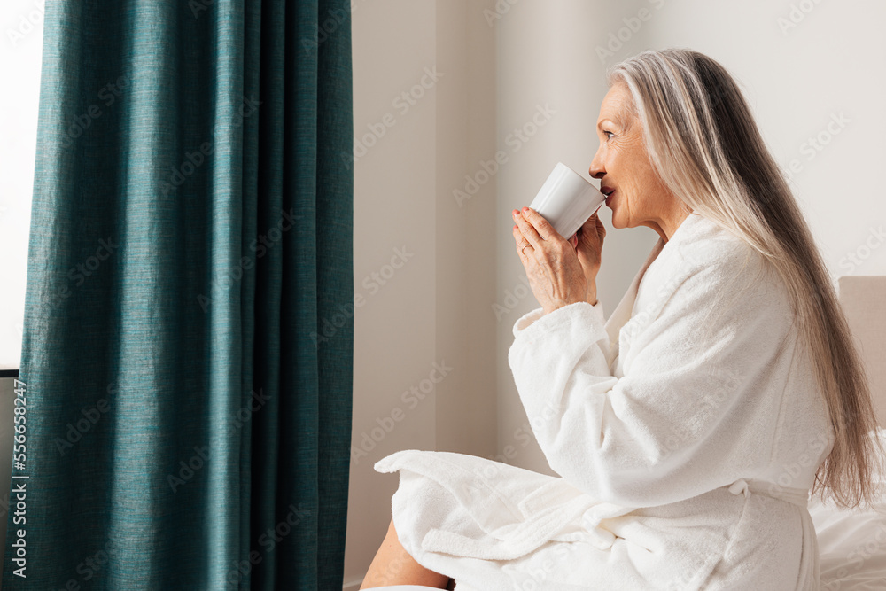 Senior woman drinking coffee in morning in bedroom. Female in bathrobe sitting on a bed looking at window. Senior woman drinking coffee in morning in bedroom. Female in bathrobe sitting on a bed looking at window.