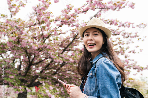 portrait of pretty asian girl wearing hat smiling at camera against blurred background of pink cherry blossom trees at springtime in mint museum in Osaka, japan