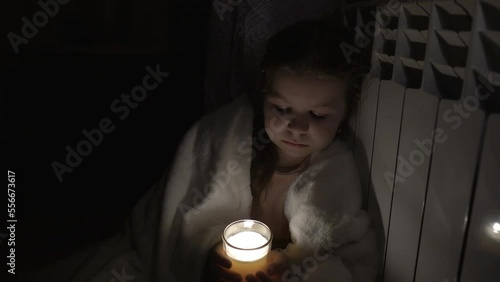 A girl in a white plaid holds a candle in a glass cup in her hands against the background of a household battery