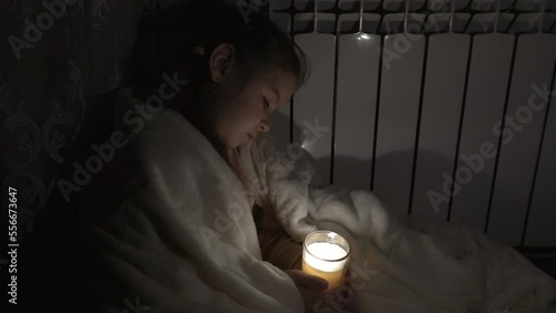 A girl in a white plaid holds a candle in a glass cup in her hands against the background of a household battery
