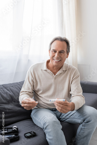 cheerful mature man with diabetes holding lancet pen and test strip while sitting on couch in living room