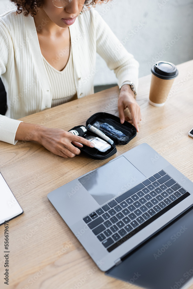 Fototapeta premium Cropped view of african american businesswoman holding diabetes kit near laptop and coffee in office