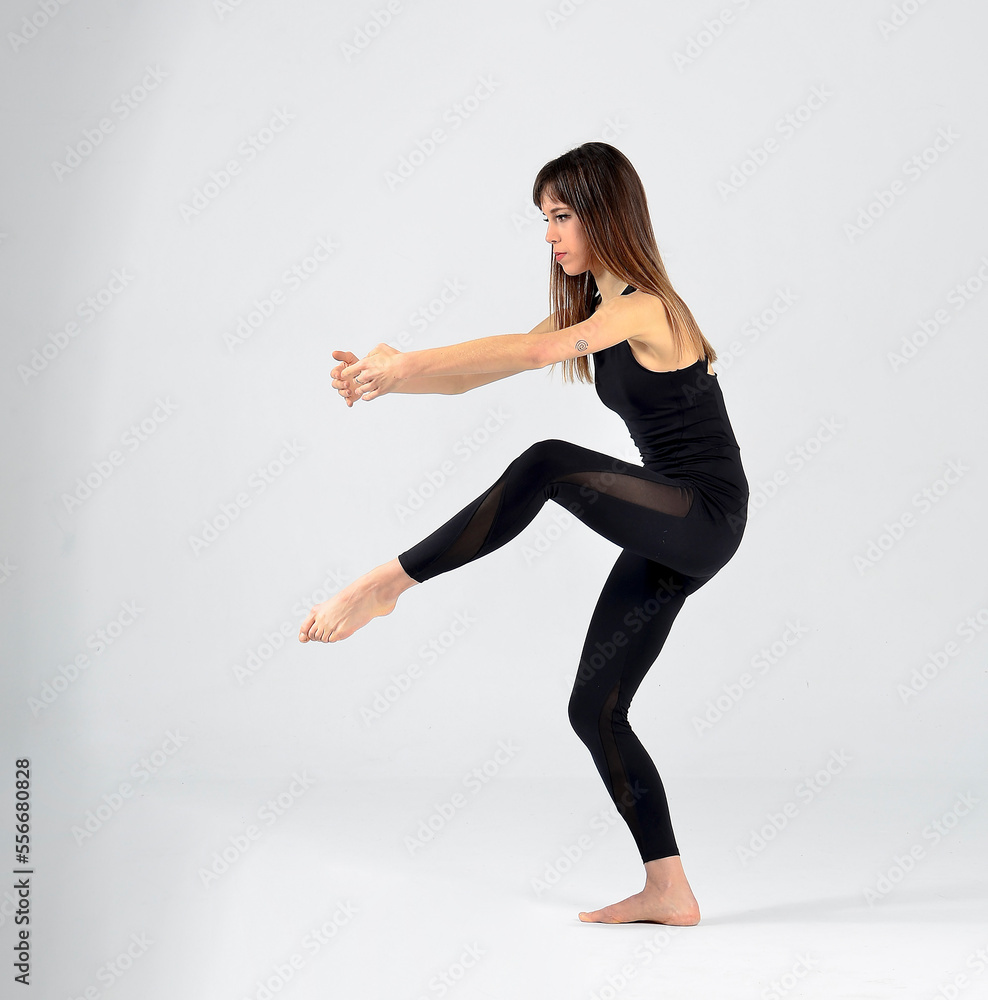 Young sporty yogi woman practicing yoga, doing Wild Thing, pose, working out, wearing sportswear, black pants and top, indoor full length, white yoga studio 