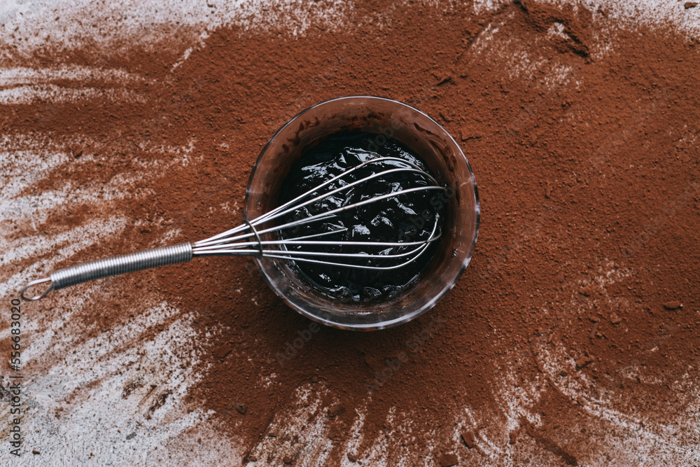 Fototapeta premium Melted chocolate and whisker on bowl against scattered cocoa powder