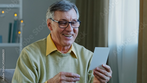 Portrait old man ecstatic grandpa with glasses sits at home indoors receives envelope letter postcard with good news offer winning victory triumph congratulations from grandchildren feeling happiness