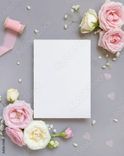 Blank paper card between pink roses and pink silk ribbons on grey top view, wedding mockup