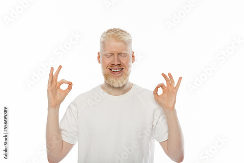 cheerful albino man with closed eyes showing ok sign with hands isolated on white