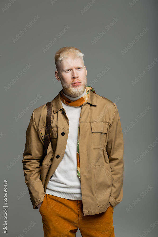 bearded albino man in stylish shirt jacket and silk scarf standing with hands in pockets isolated on grey