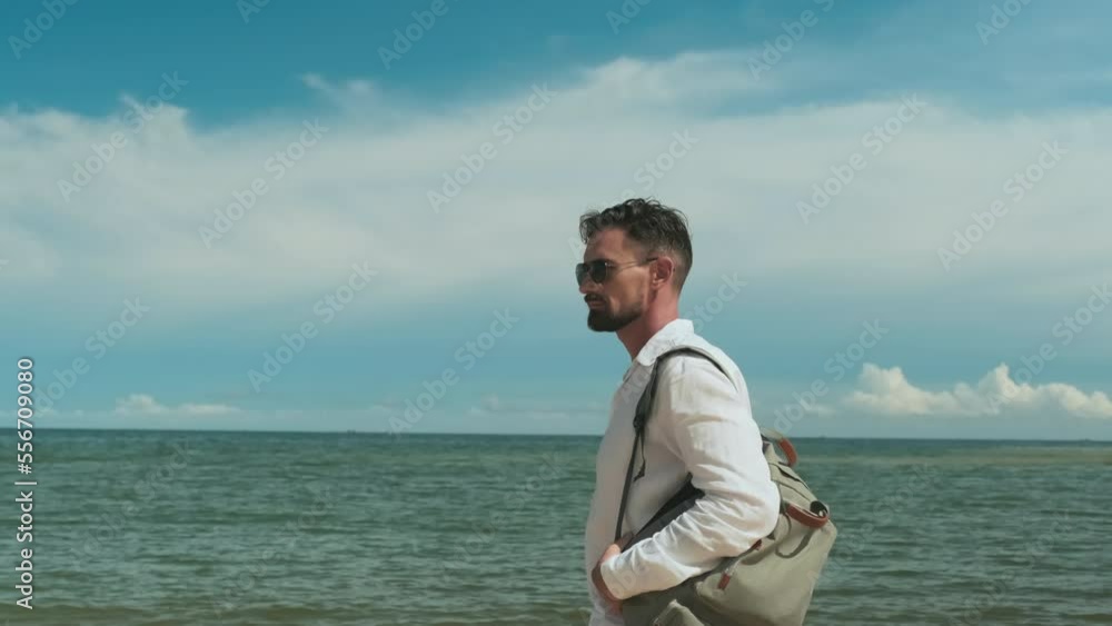 Young restful man with backpack on shoulder moving along seaside against cloudy sky on hot summer day at tropical resort and looking at camera while enjoying vacation Young restful man with backpack on shoulder moving along seaside against cloudy sky on hot summer day at tropical resort and looking at camera while enjoying vacation