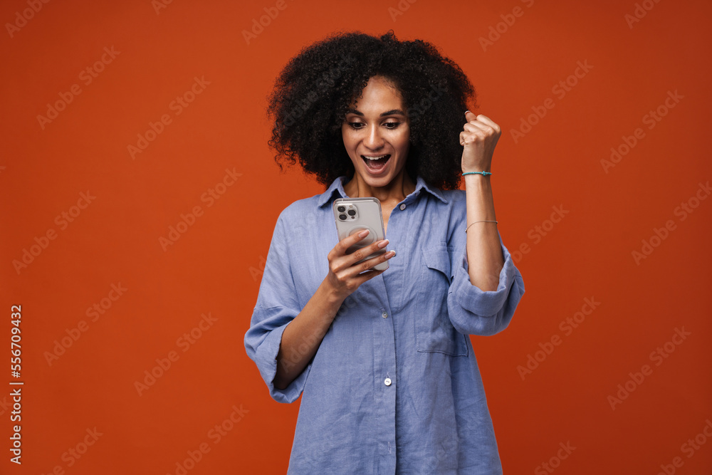 Smiling african american woman using mobile phone and showing winner gesture isolated over red wall Smiling african american woman using mobile phone and showing winner gesture isolated over red wall