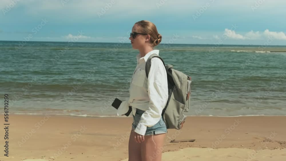 Carefree girl with backpack walking along coastline, stopping to text message in smartphone and continuing her way through tropical beach on hot sunny morning while enjoying vacation Carefree girl with backpack walking along coastline, stopping to text message in smartphone and continuing her way through tropical beach on hot sunny morning while enjoying vacation
