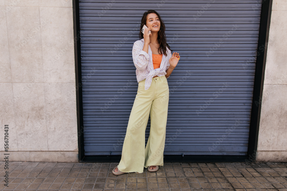 In full growth of cool young caucasian woman speaking on her smartphone outdoor. Brunette wears shirt, top and casual jeans. Concept of smart devices. In full growth of cool young caucasian woman speaking on her smartphone outdoor. Brunette wears shirt, top and casual jeans. Concept of smart devices.
