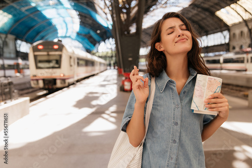 Beautiful young caucasian girl stands at railway station with closed eyes crossed her fingers to luck. Brunette wears denim jeans jacket. Trip concept.