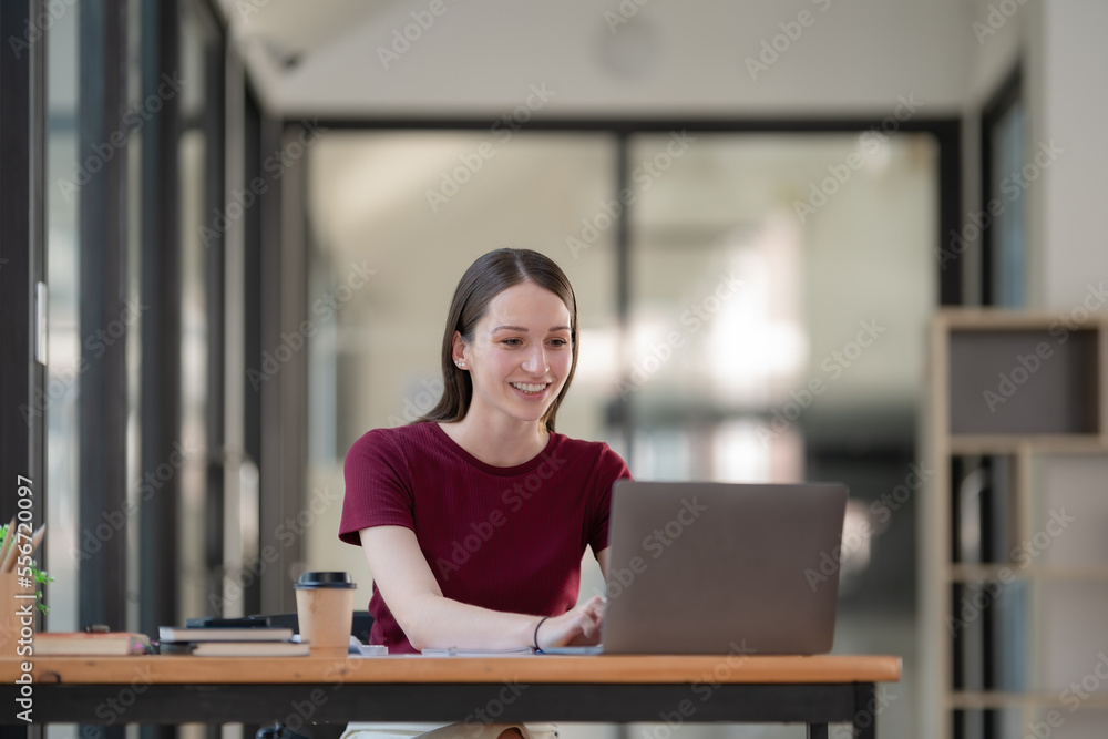 Concentrated at work. Confident young woman in smart casual wear working on a laptop while sitting near a window in a creative office or cafe Concentrated at work. Confident young woman in smart casual wear working on a laptop while sitting near a window in a creative office or cafe