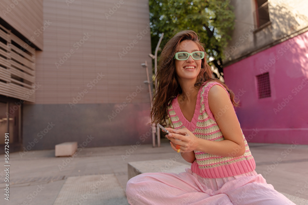 Positive young caucasian girl in sunglasses looks away sitting outdoor with smartphone. Woman wears vest and casual pants. Happy holiday, device use concept.