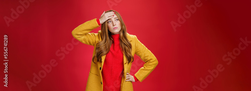 Girl having feaver touching forehead with palm looking displeased and bothered left as thinking she got cold standing in yellow coat, realising she sick or ill over red background, feeling tired photo