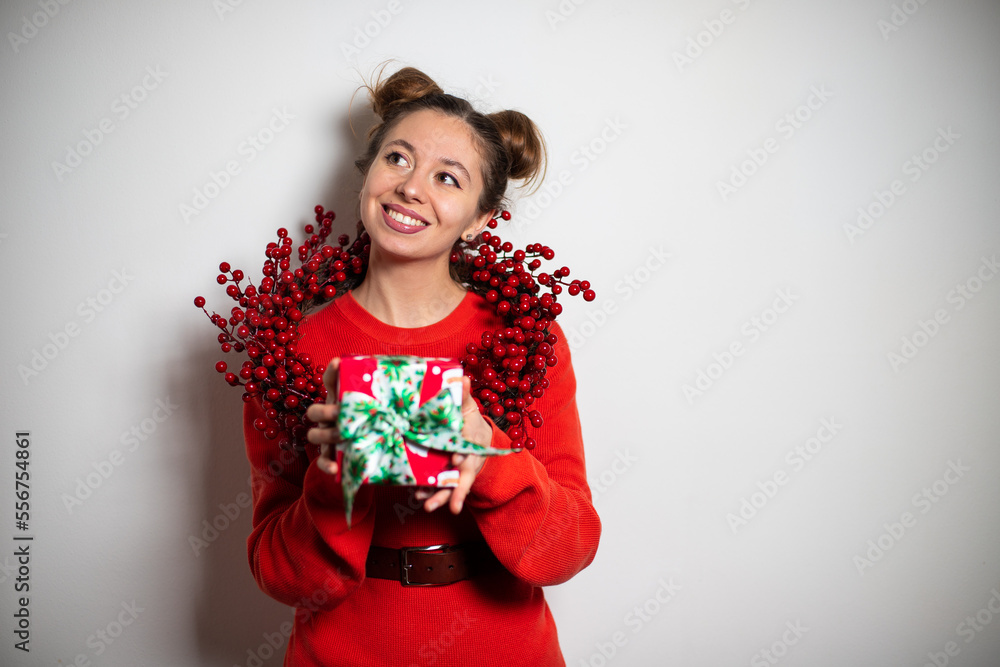 Portrait of young cute brunette girl with two bundles, red sweater, red Christmas garland smiling widely holding the red green present in hands Portrait of young cute brunette girl with two bundles, red sweater, red Christmas garland smiling widely holding the red green present in hands