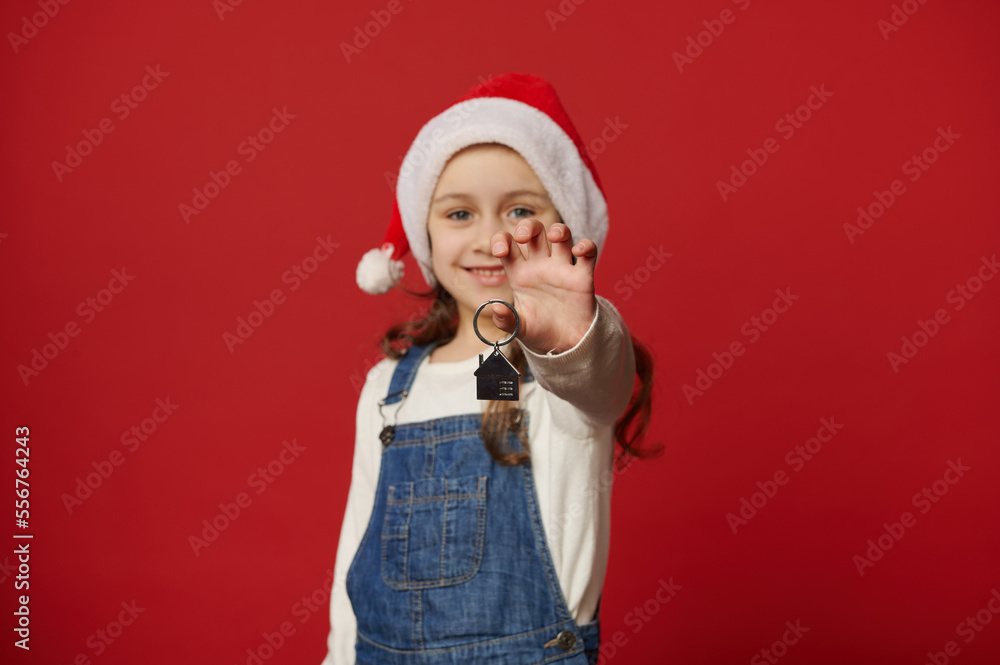 Details: keychain in the shape of new house, in hands of adorable smiling little girl in Santa hat and denim overalls. New apartment, private residence - best New Year present. Real estate concept Details: keychain in the shape of new house, in hands of adorable smiling little girl in Santa hat and denim overalls. New apartment, private residence - best New Year present. Real estate concept