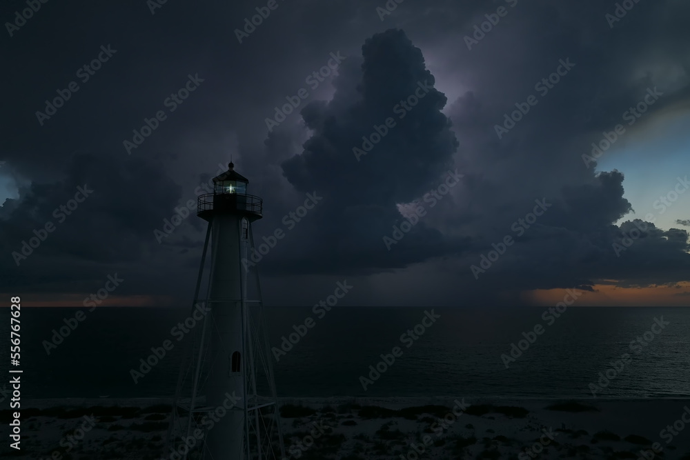 White tall lighthouse on sea shore with blinking light at stormy night for commercial vessels navigation. Thunderstorm with lightnings over ocean water posing danger for ships