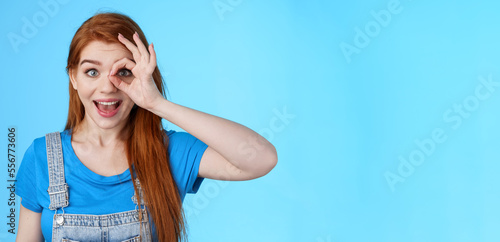 Dreamy curious attractive cheerful redhead woman surprised, look fascinated camera look through fingers okay sign, stand amused, gaze admiration blue background, check out promo