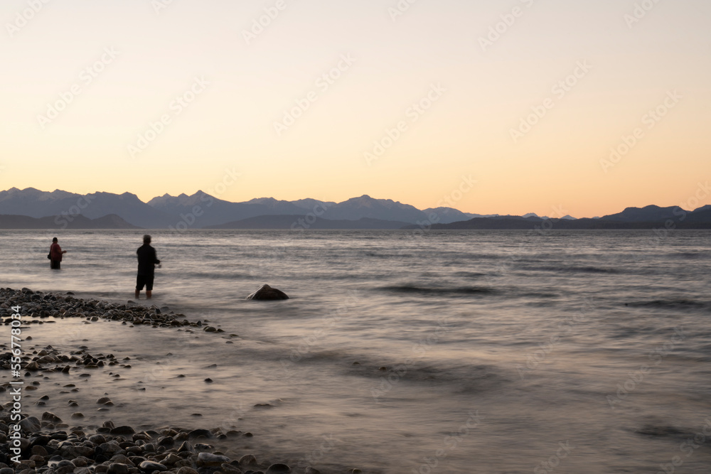Night shot. View of the water blurred effect and blurred fishermen dark silhouette, fishing at nightfall from the coast. The rocky shore and mountains in the horizon with a beautiful dusk light.