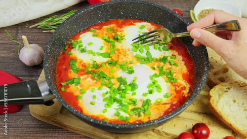 Person eating shakshuka, tomato eggs served in pan on wooden table. hands cutting poached eggs in tomatoes with fork, spicy shakshouka, traditional Israeli breakfast, slow motion video, 4k fottage photo