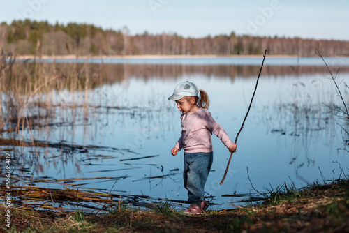 A three-year-old child walks along the shore of the lake in the spring, on a sunny day.