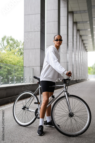 Fototapeta Naklejka Na Ścianę i Meble -  Fit and stylish african american woman standing near bicycle on road 
