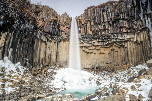 Cascata Svartifoss, torrente d'acqua che cade da un salto di rocce basaltiche. Sassi sul letto del torrente, ghiaccio alla base della cascata, stalattiti di ghiaccio photo