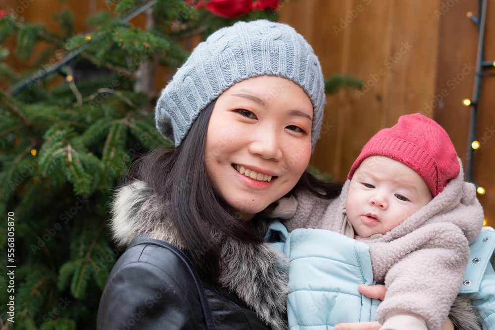 young happy and attractive Asian Korean woman holding baby girl in her arms in front of Christmas tree walking on Winter street market in mother and daughter love concept young happy and attractive Asian Korean woman holding baby girl in her arms in front of Christmas tree walking on Winter street market in mother and daughter love concept