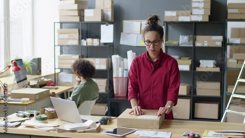 Woman attaching paper label to parcel box while going to ship online store order in delivery service office